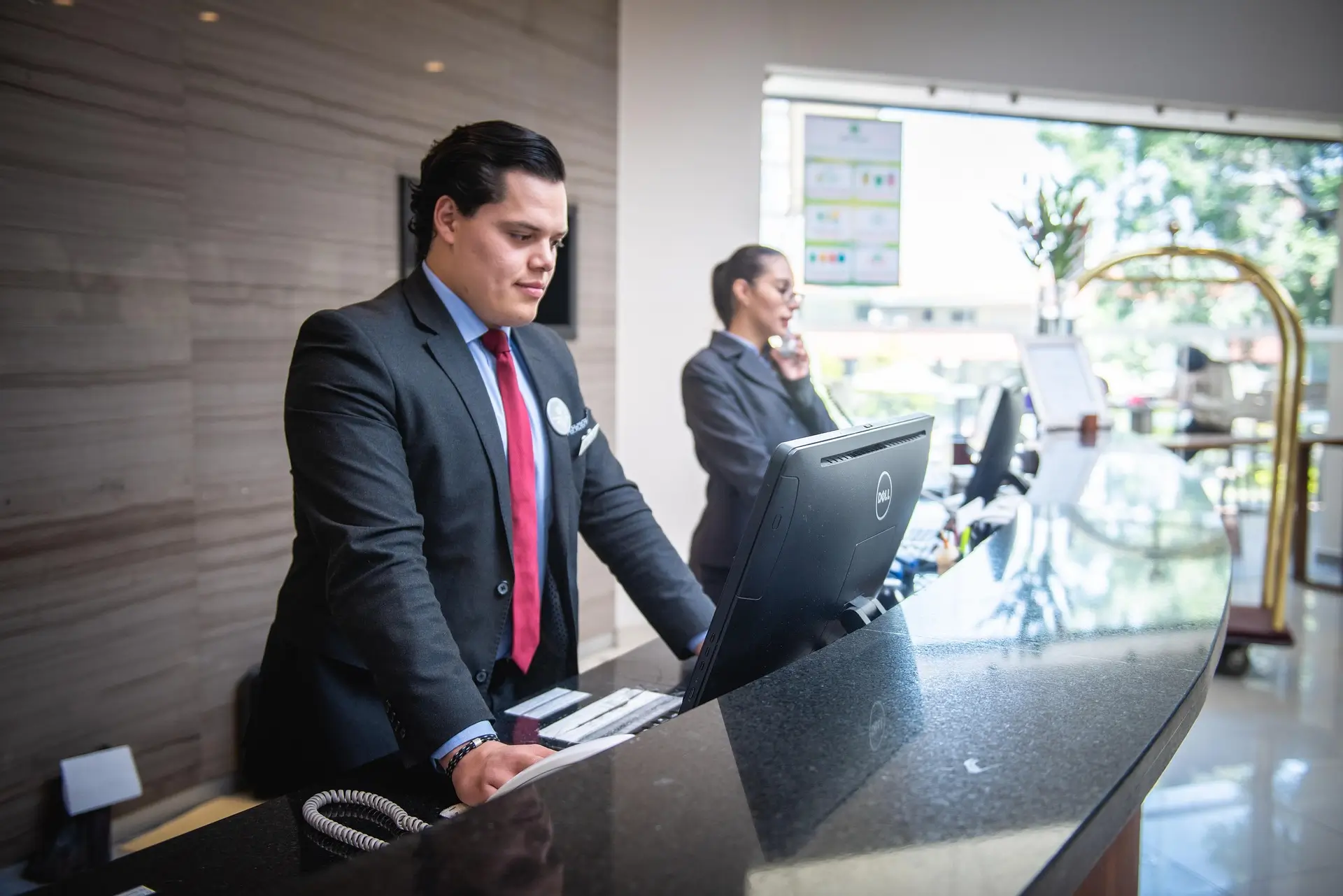Recepcionista de hotel trabalhando no computador do balcão da recepção, com uma colega ao telefone ao fundo